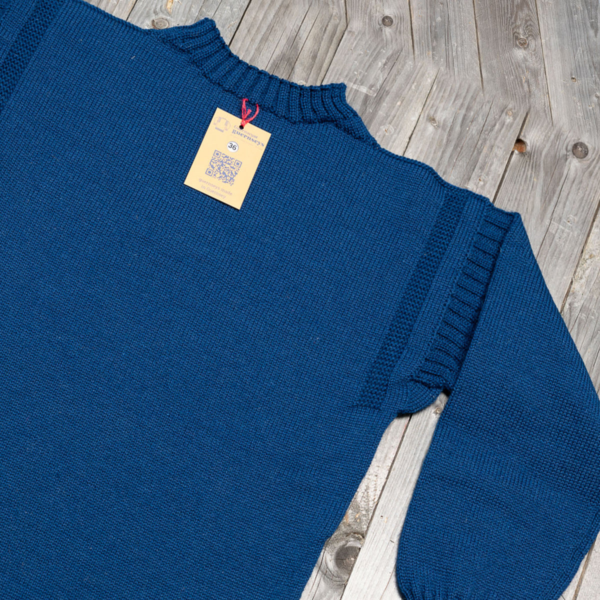 Traditional GG Blue wool guernsey sweater featuring a ribbed shoulder detail and a size 36 tag on a rustic wooden background.