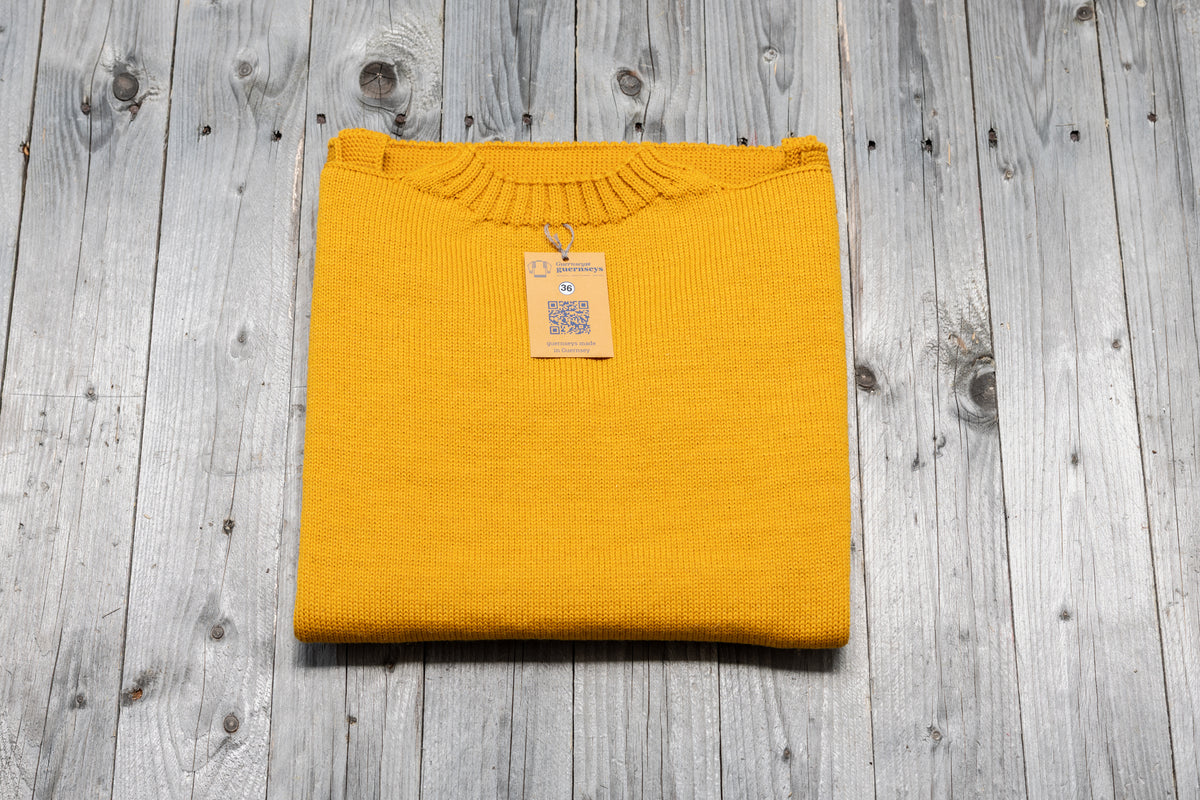 Folded mustard yellow pure wool traditional guernsey sweater with a size 36 tag on a rustic grey wooden background.