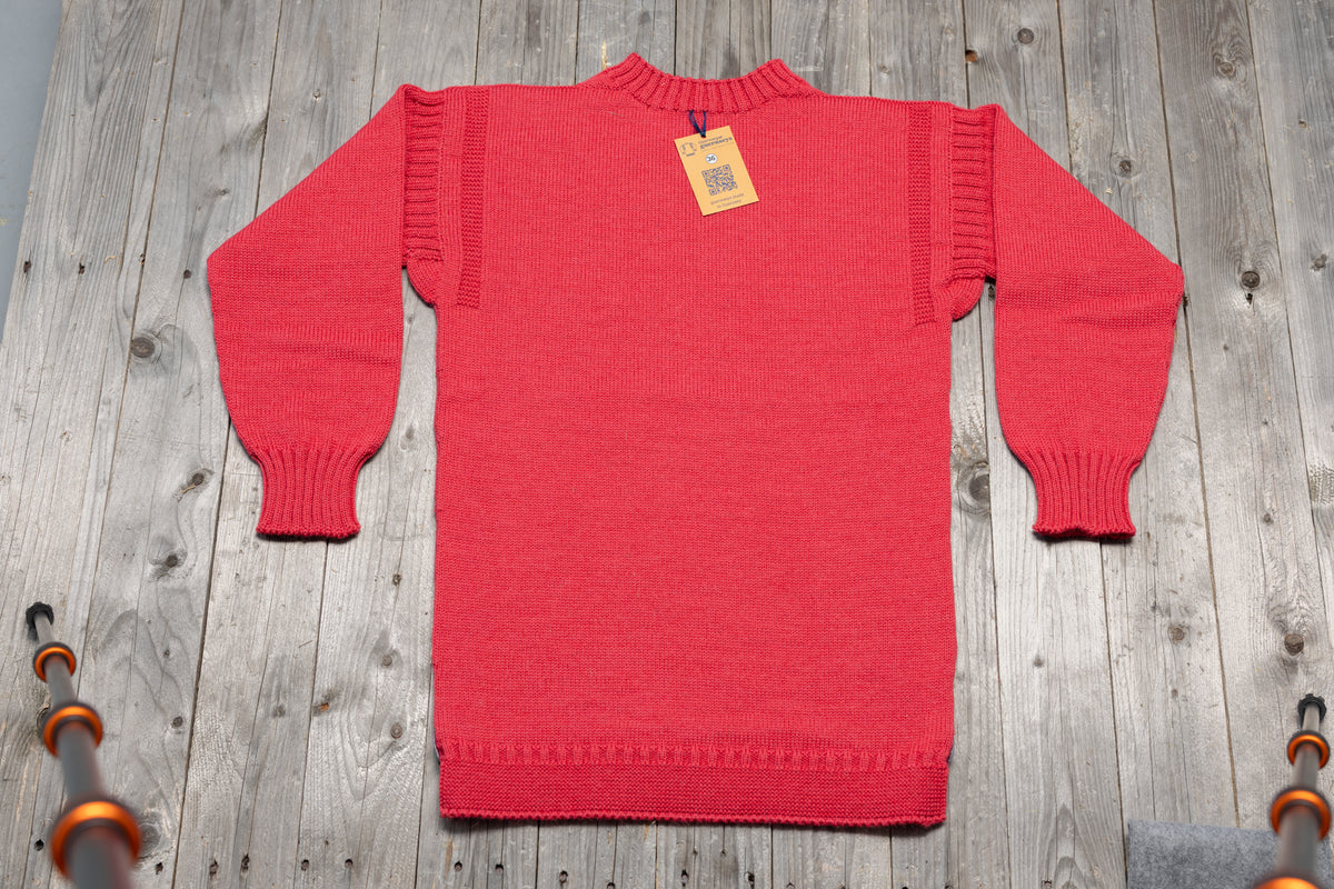 Traditional Lily pink wool guernsey sweater with ribbed cuffs and neckline, laid flat on a rustic wooden background.