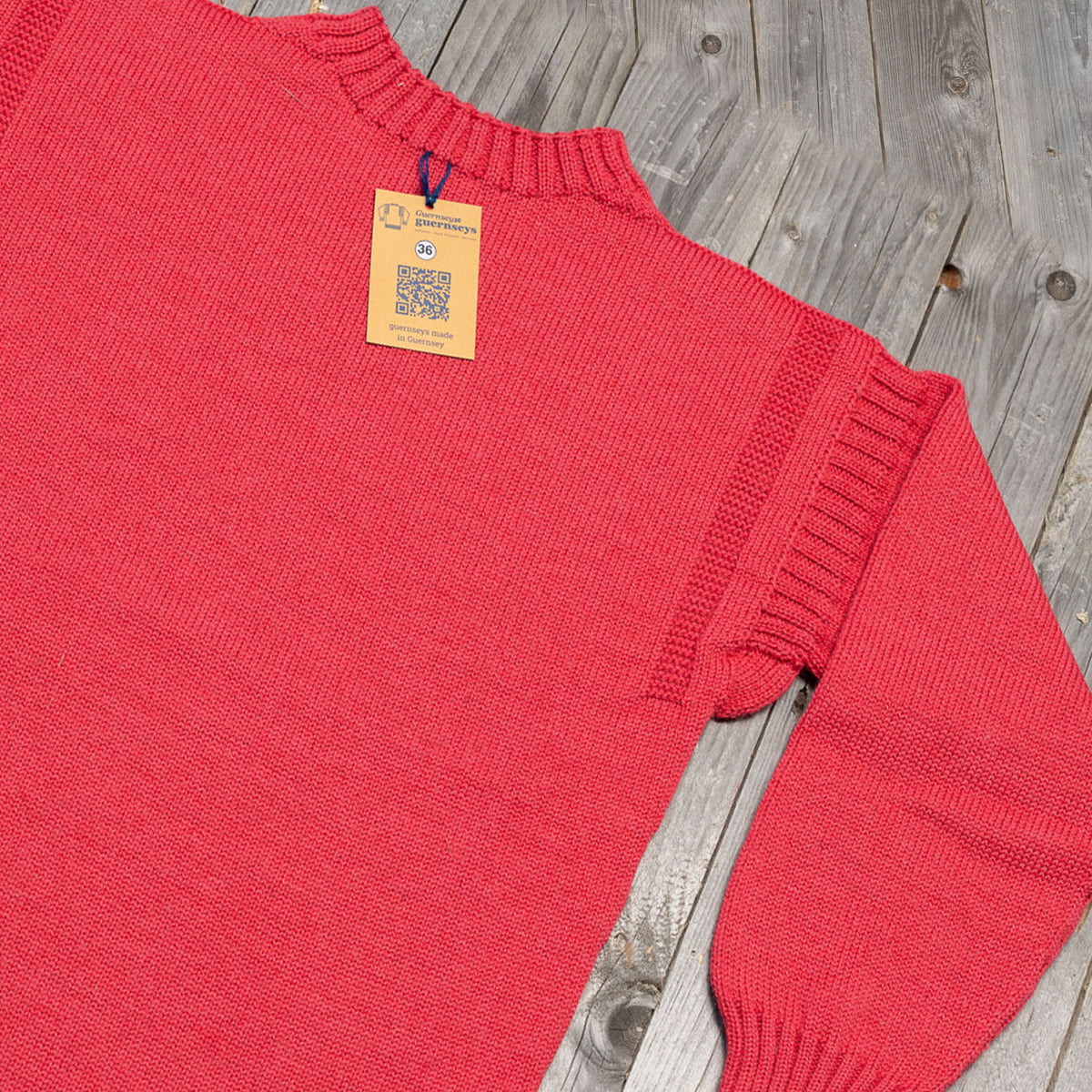 Traditional Lily pink wool guernsey sweater with ribbed collar and shoulder detailing, featuring a size 36 tag on wooden planks.