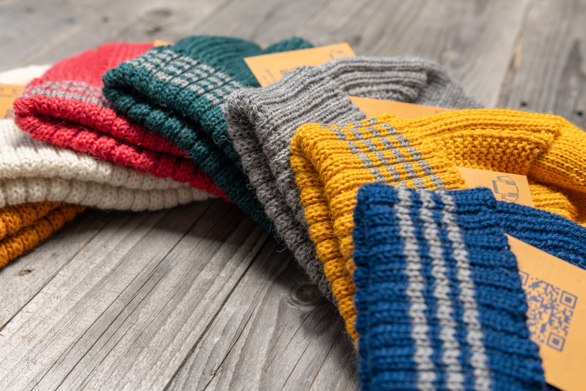 Stack of colorful, ribbed wool Hanois beanies featuring reflective bands on the turn-back cuffs, resting on a wooden surface.